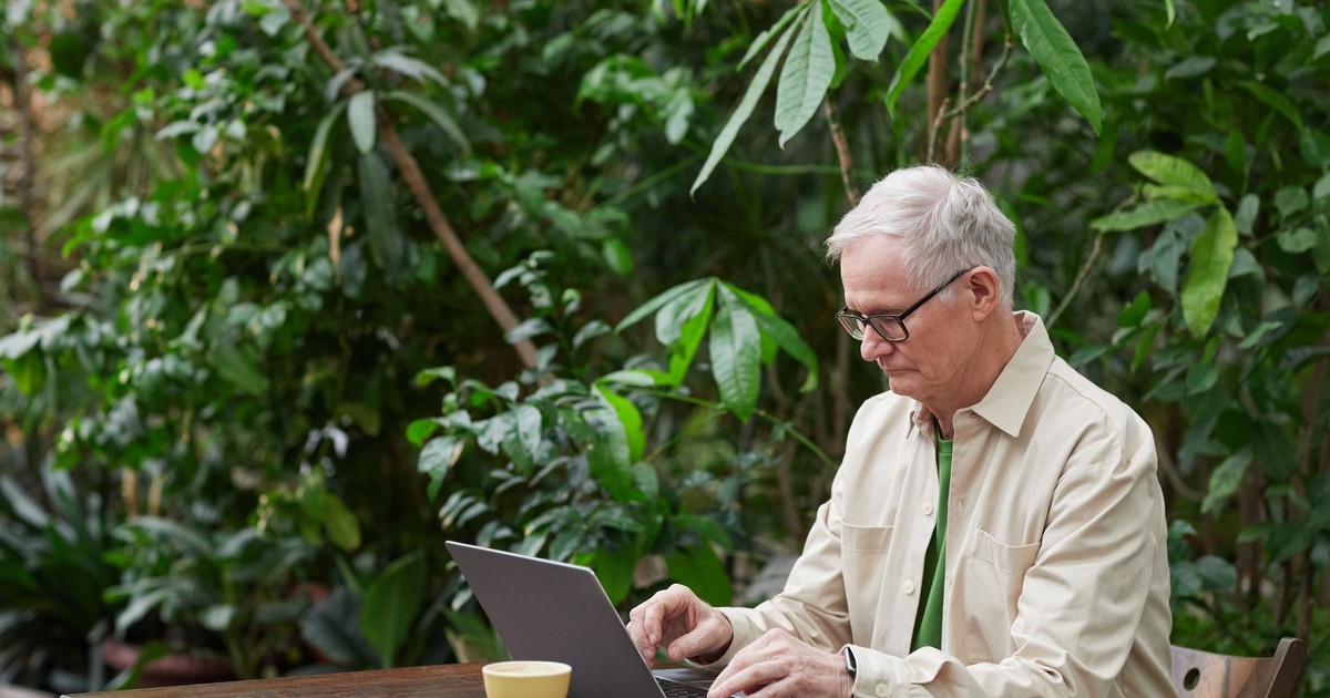 Older man using a laptop in a green garden setting, illustrating remote work and technology usage.