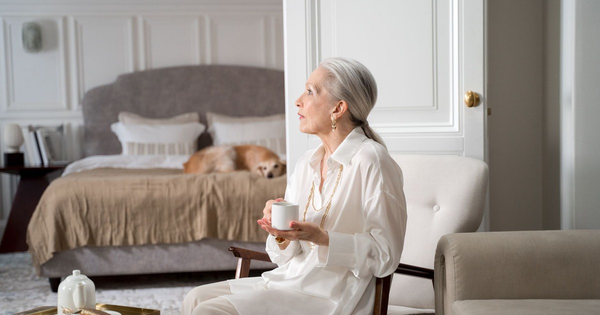 Senior woman with gray hair sipping coffee in a cozy living room with a dog resting on the bed.