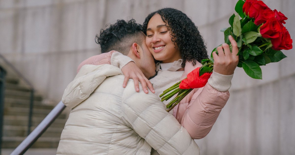 Anonymous enamored young ethnic man in warm clothes hugging cheerful girlfriend smiling with closed eyes after receiving bouquet of fresh red roses