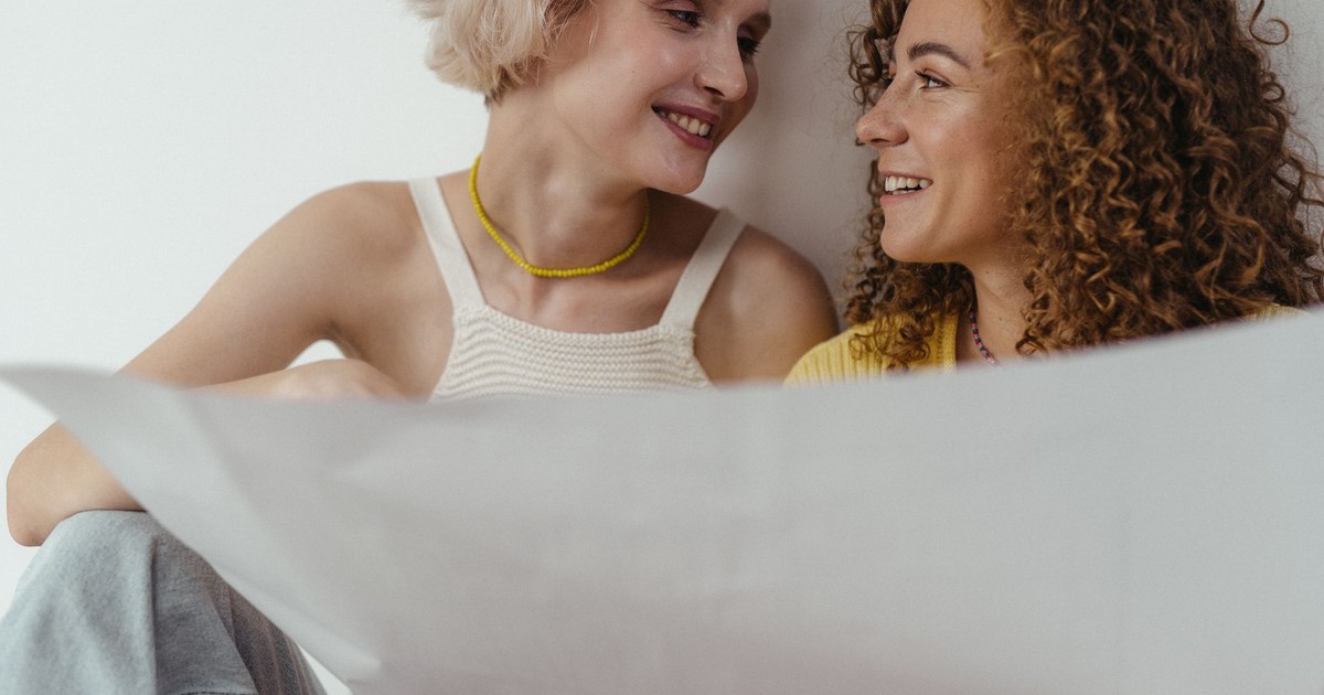 LGBTQ+ couple smiling and sharing a moment while holding a blueprint indoors.