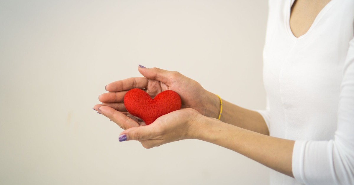 Unrecognizable lady wearing white outfit standing on white background with decorative small red heart in hands in light room inside