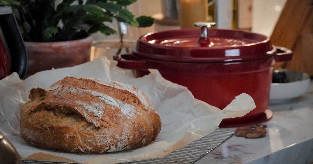 Close-up of a rustic loaf of bread on a kitchen counter with a red Dutch oven nearby.