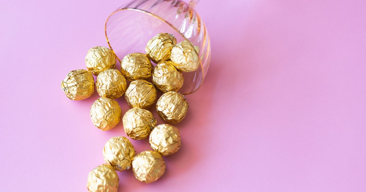 Elegant arrangement of gold foil wrapped chocolates in a cocktail glass on a pink background.
