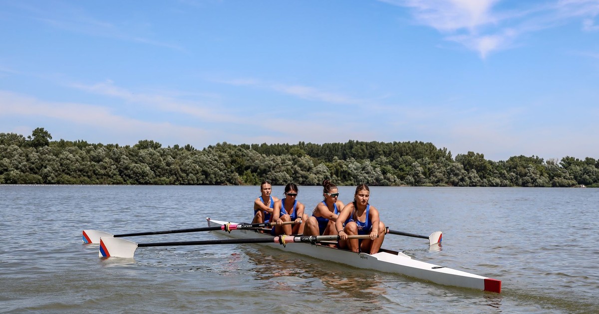 Four women in a rowing boat practicing on a calm river under a clear blue sky.