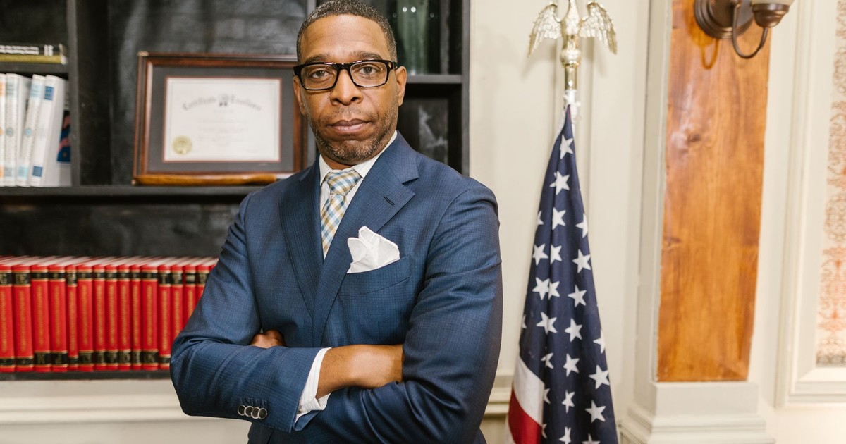 Portrait of a confident lawyer in office attire, standing with arms crossed in front of a USA flag.