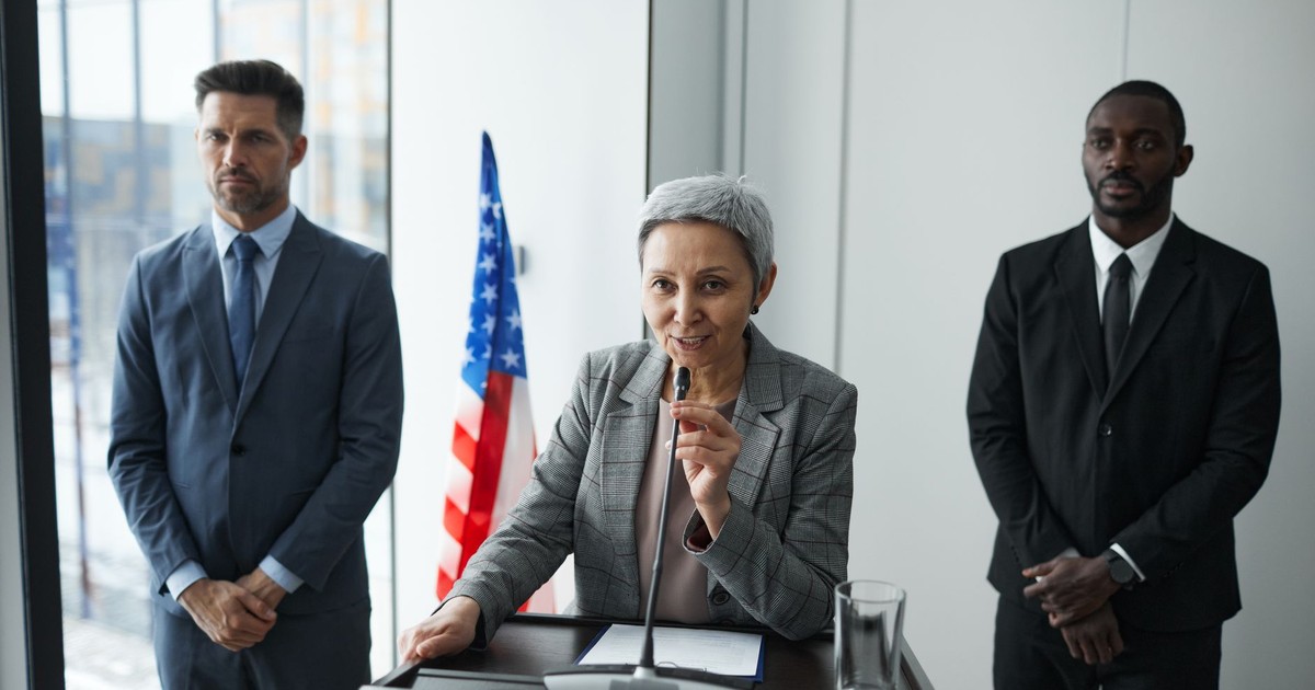 A female politician delivers a speech with bodyguards and an American flag in the background.