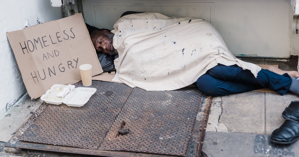 Street scene depicting a homeless man sleeping with a placard reading 'Homeless and Hungry'.