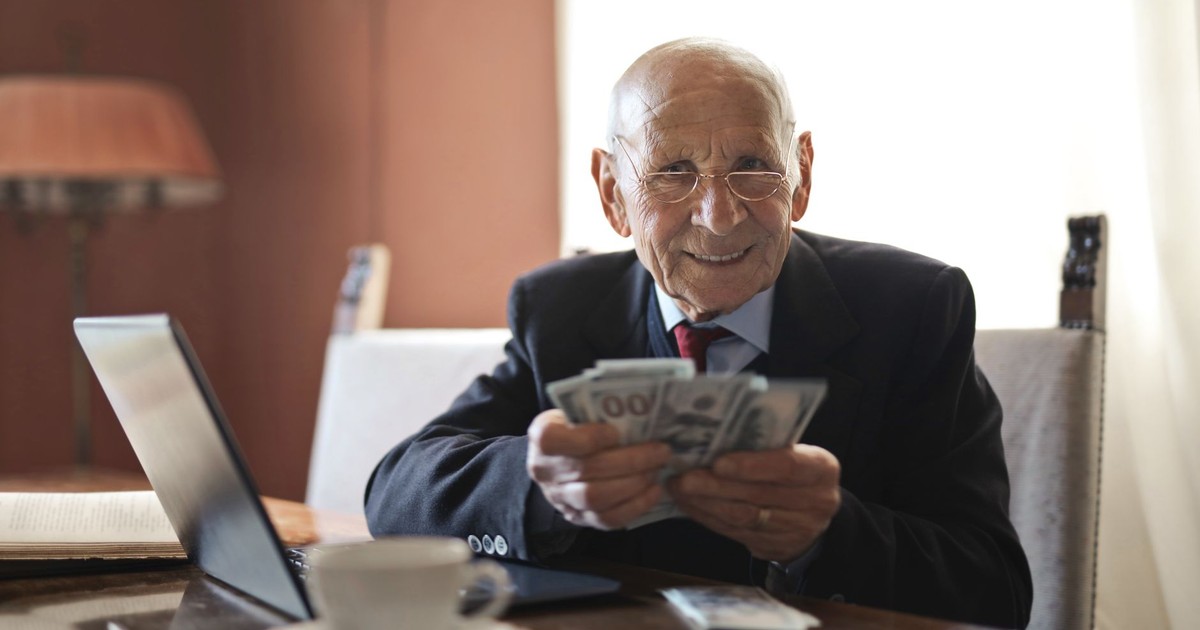 Positive senior businessman in formal suit and eyeglasses holding money in hands while sitting at table near laptop and looking at camera