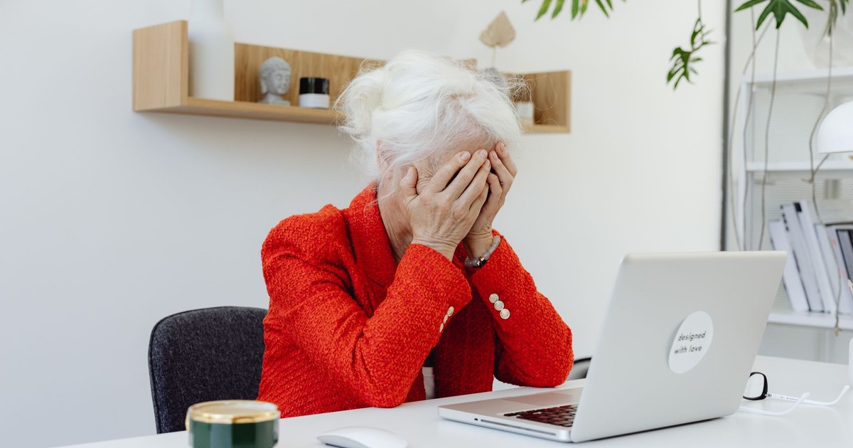 Elderly woman in red jacket looking frustrated at laptop in home office setting.