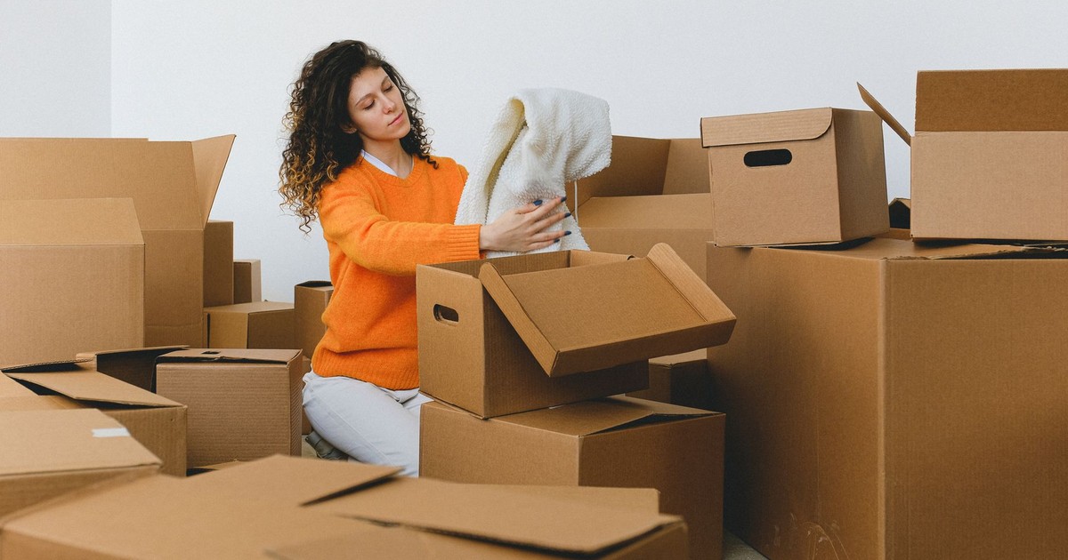 Calm young relocating woman with curly dark hair in stylish sweater packing belongings into pile of cardboard boxes sitting on floor before moving into new apartment