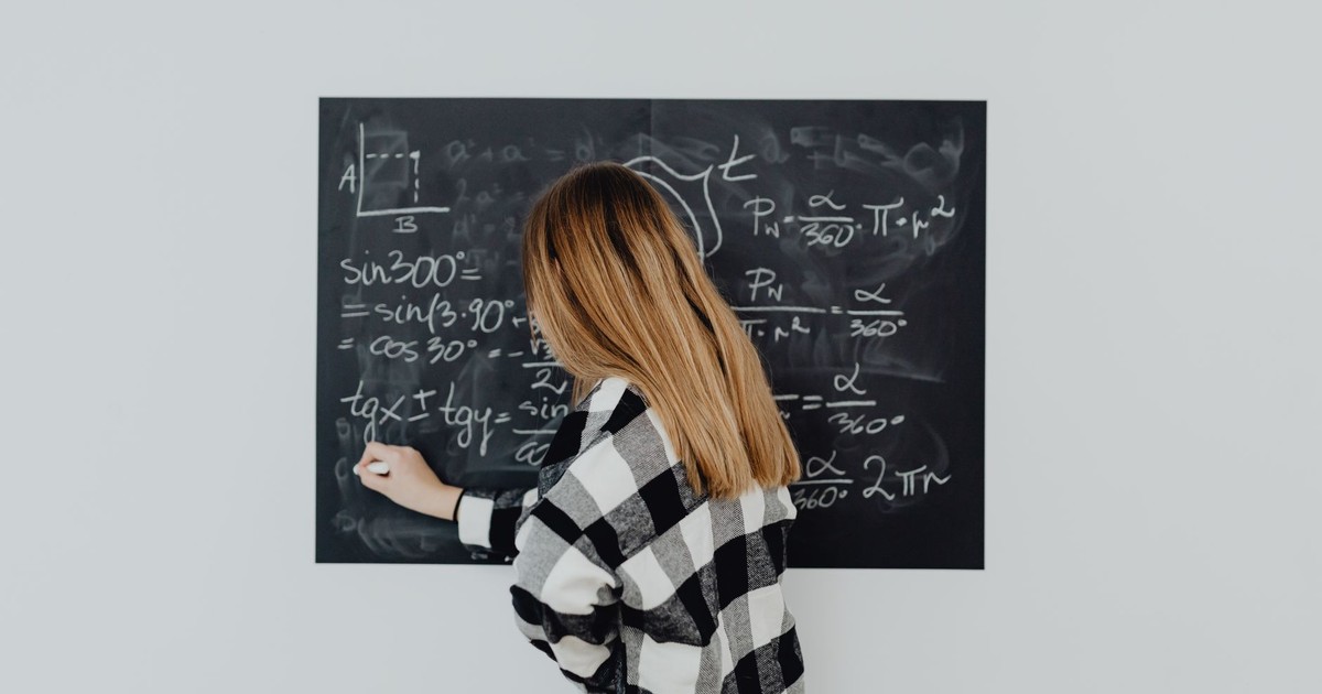 Teen girl in plaid shirt solving math equations on a blackboard, focusing on trigonometry.