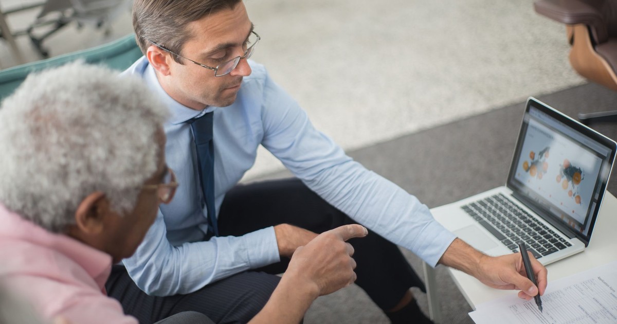 Two men in a business meeting reviewing financial documents and discussing data on a laptop.
