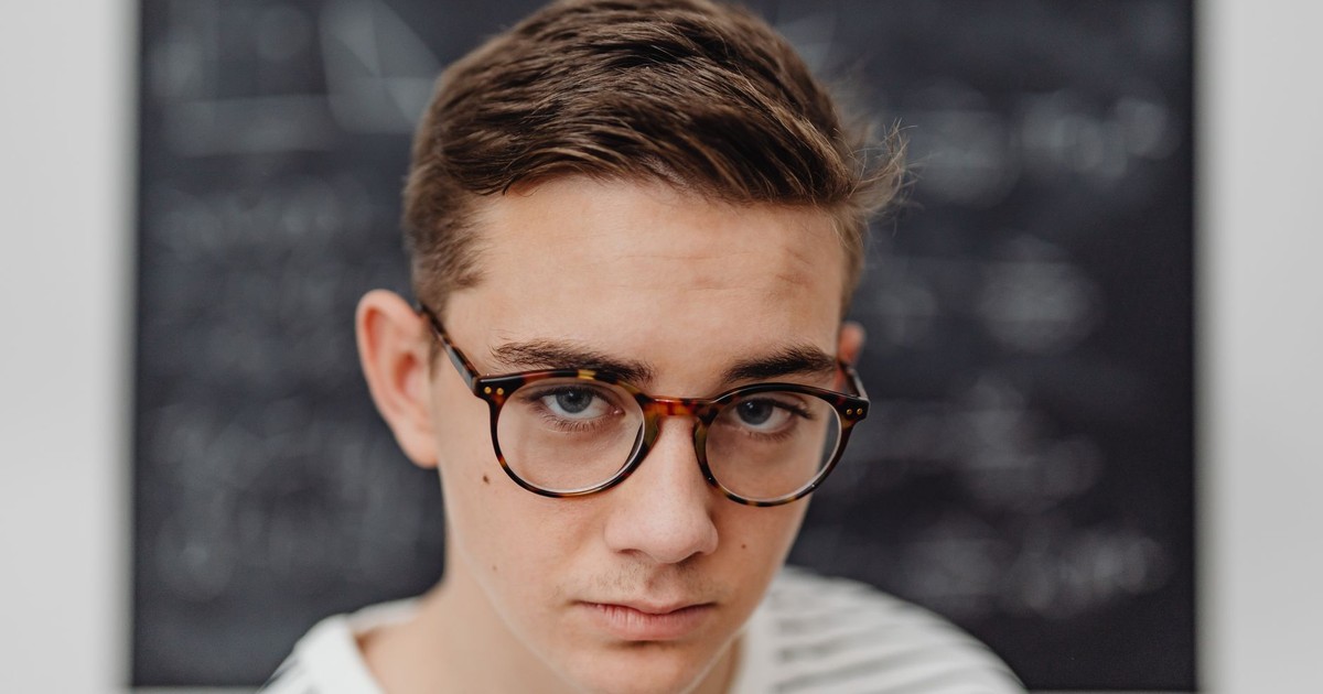 Teenage male student wearing glasses stands in front of a school blackboard.