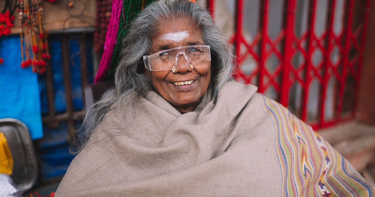 Elderly woman with glasses and traditional shawl smiling at a Varanasi market stall.