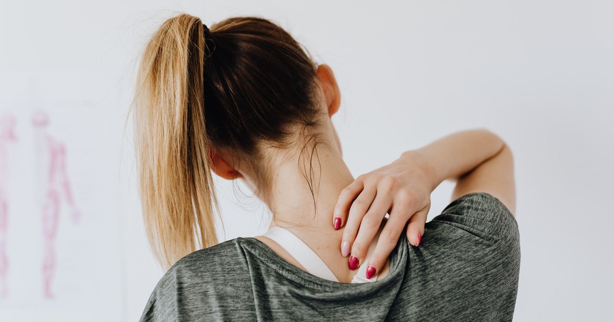 Back view of slim blond female in casual wear with manicure indicating pain spot on back while sitting in front of white wall with paper drawing in clinic