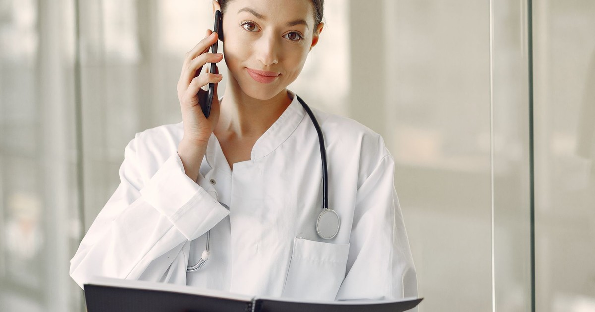 Crop ethnic female doctor in medical uniform with stethoscope and notebook talking on cellphone while standing in clinic corridor