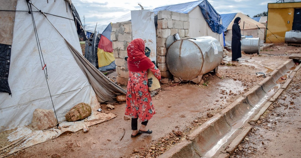 Full body of poor women in hijabs and headscarves near weathered buildings and camps in village under cloudy sky