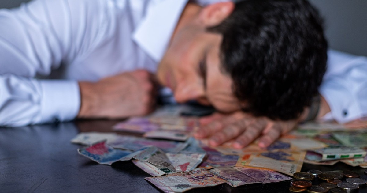 A man in a white shirt rests his head on a table covered with Argentine pesos and coins.