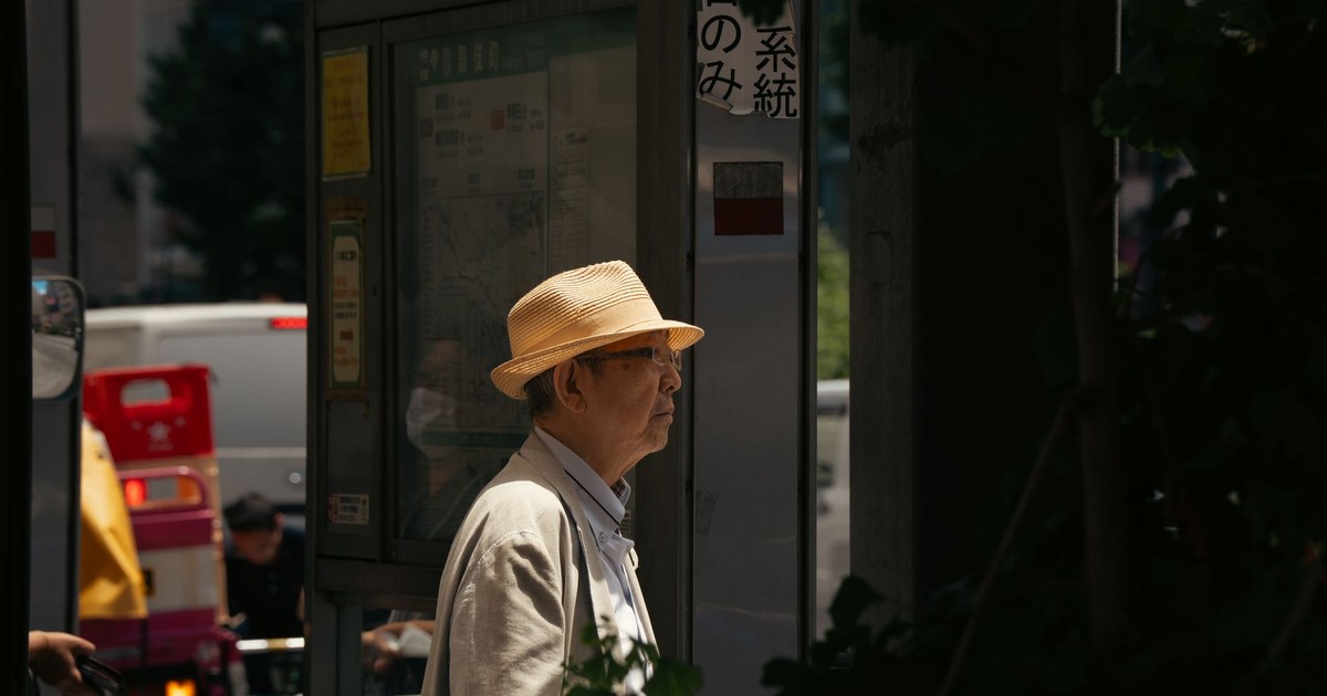 Elderly man in a straw hat stands at a bus stop in a shaded Tokyo street.