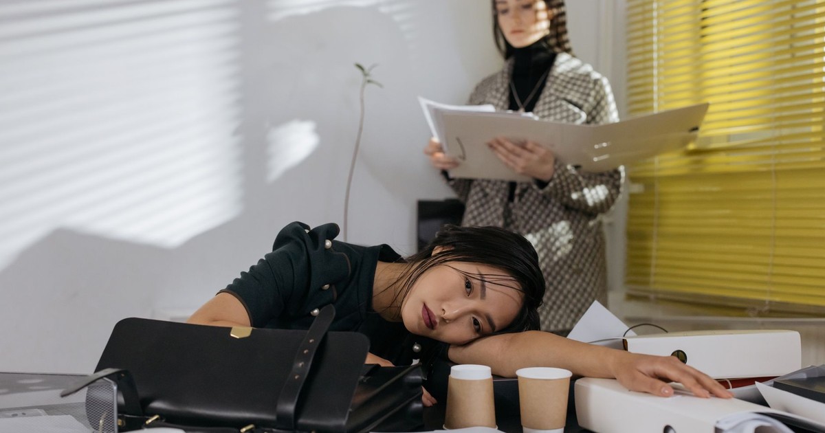 Exhausted employee resting on desk surrounded by documents, colleague in background.