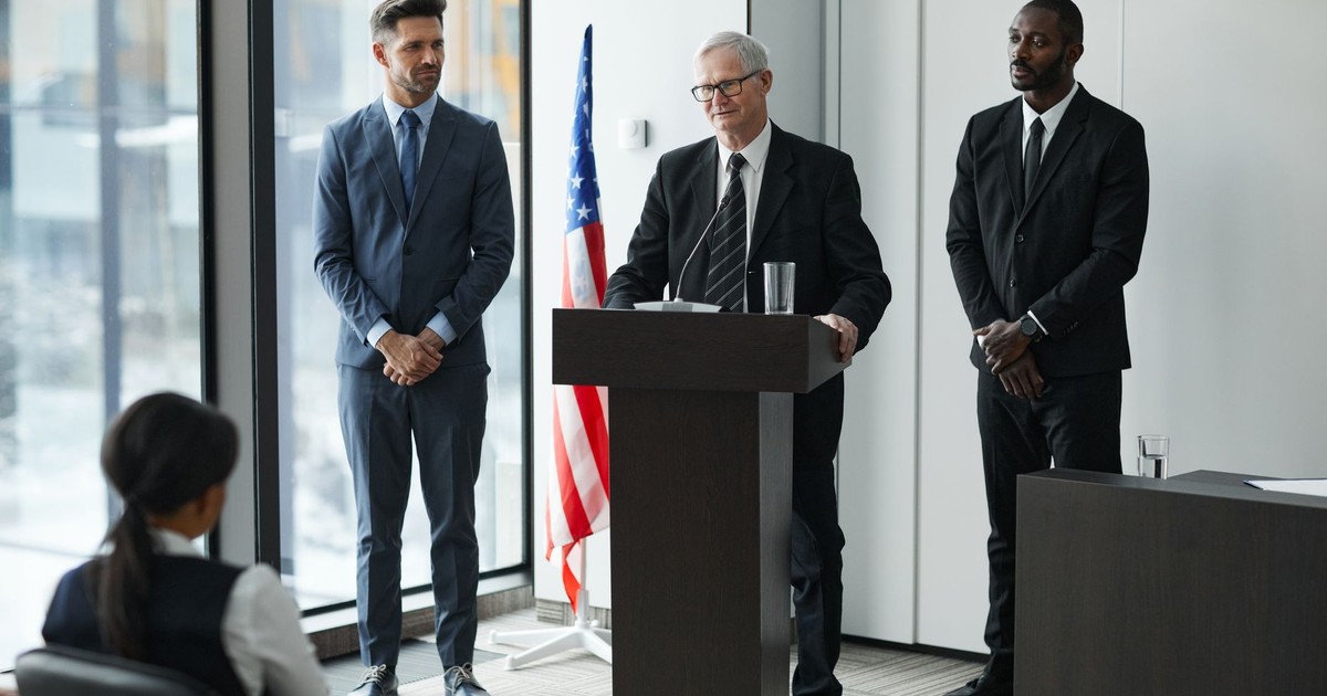 Officials delivering a political speech in a modern conference room with an American flag.