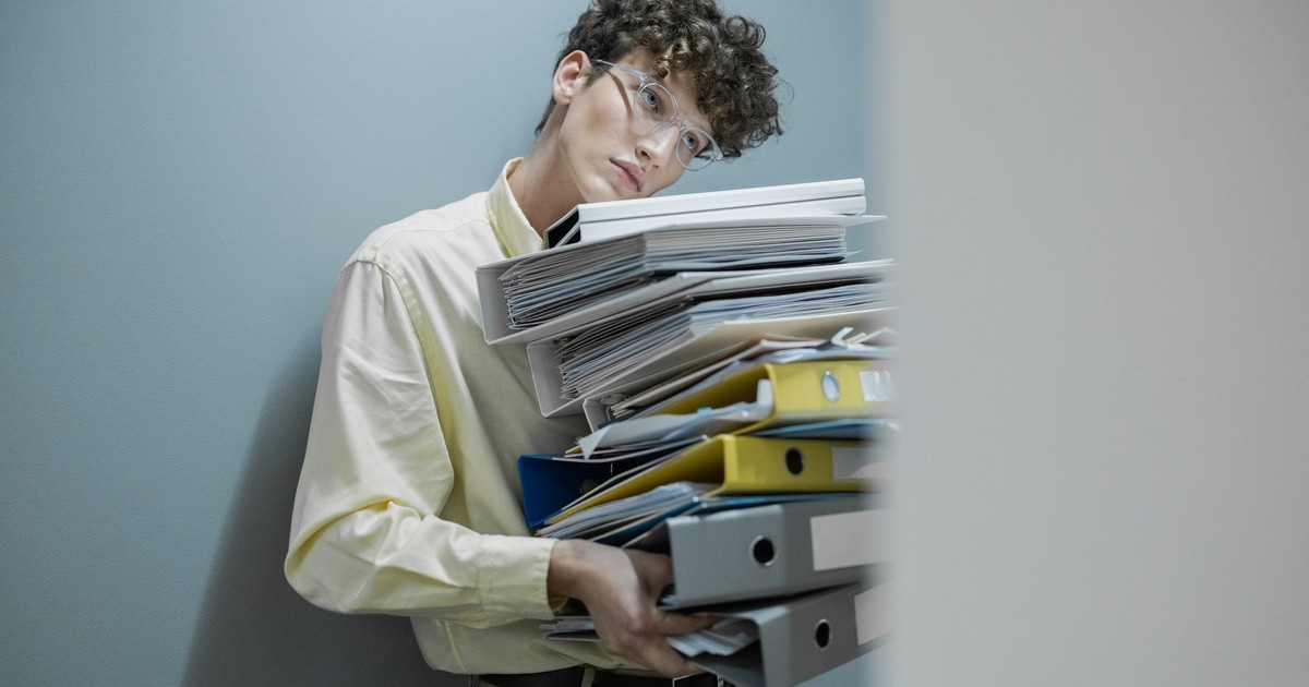 Young man overwhelmed with a stack of folders symbolizes workplace stress.