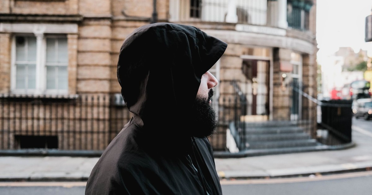 Bearded man in a hoodie walks past urban architecture on a city street corner.