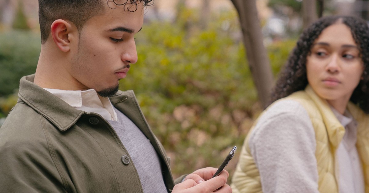 Side view of Latin American male surfing on phone while girlfriend looking seriously and sitting in park near trees and bushes in daytime