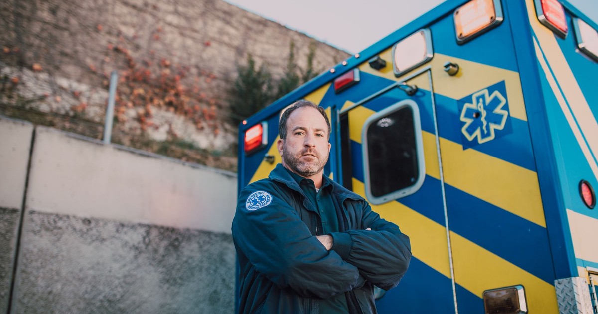 Male paramedic standing confidently beside a blue and yellow ambulance outdoors.