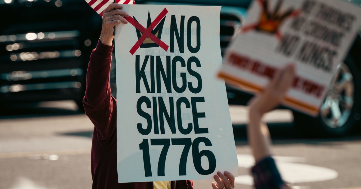 Protest sign displaying 'No Kings Since 1776' held alongside a small US flag, emphasizing democratic sentiments.