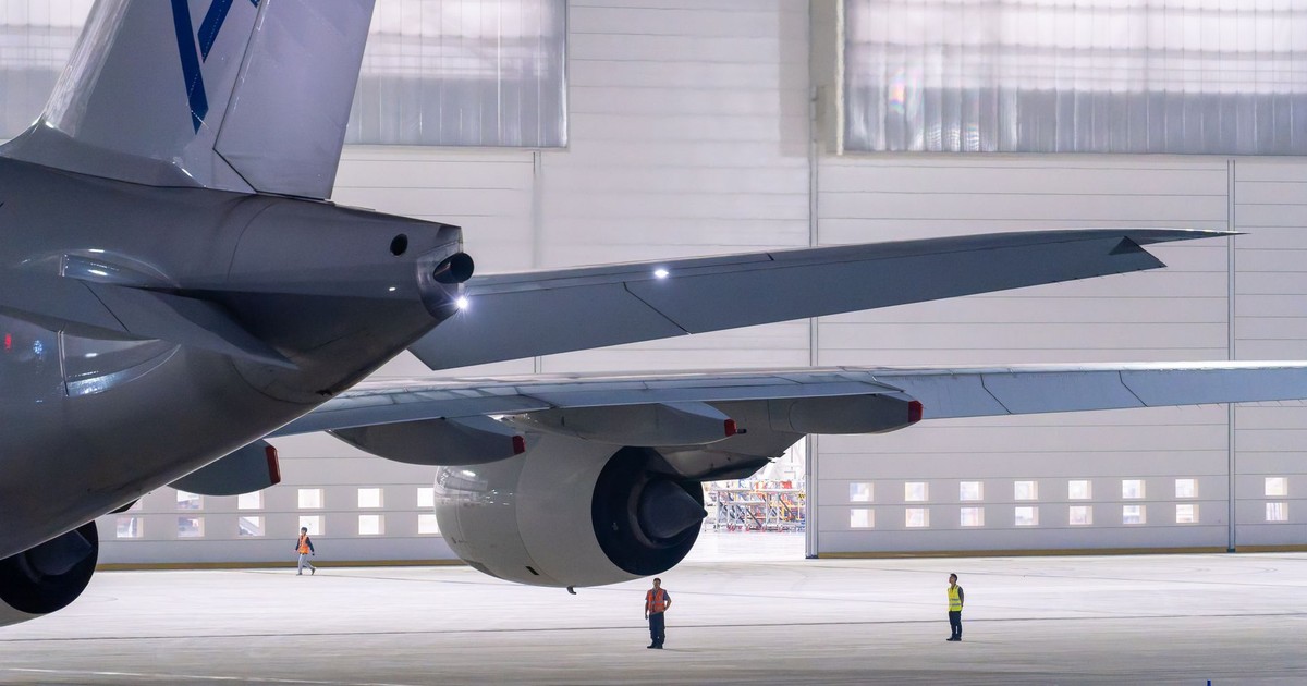 Large commercial airplane parked in an illuminated airport hangar with maintenance staff visible.