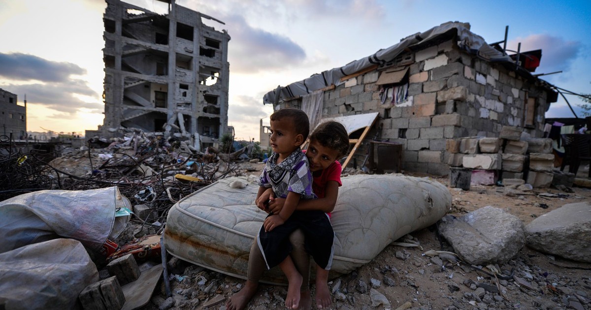 Two children sit on a mattress amidst the ruins of a building in an urban decay setting during early twilight.