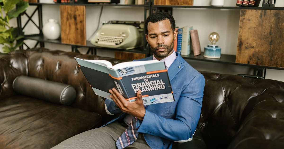 A man in a blue blazer sits on a leather couch reading a financial planning book.