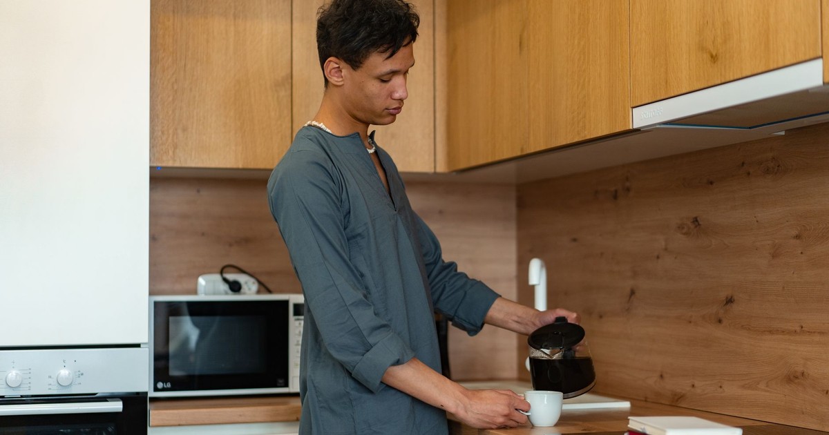 A man in a kaftan pours coffee into a white mug in a stylish wooden kitchen setting.