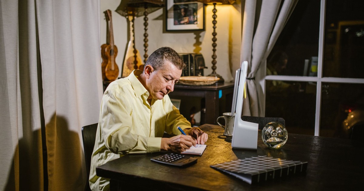 Senior man wearing yellow shirt writing notes at desk with lamp, calculator, and glass sphere nearby.