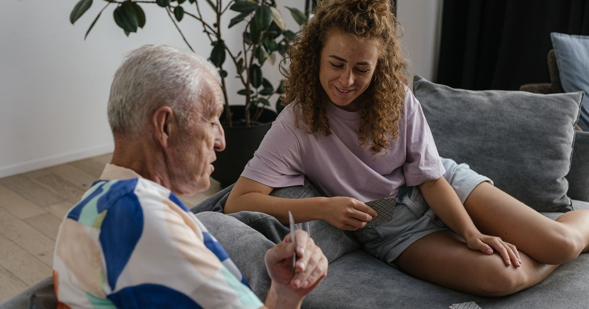 A senior adult and young woman share a joyful moment playing cards indoors on a cozy couch.