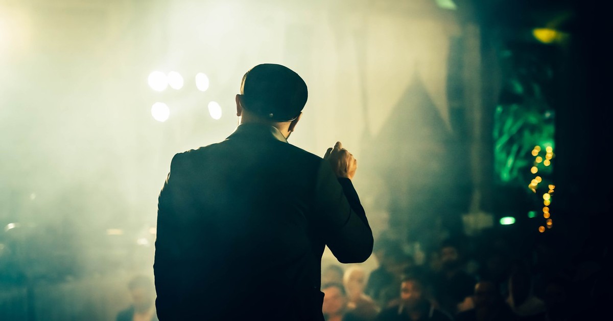 A man seen from the back speaking to an audience at an atmospheric entertainment venue.