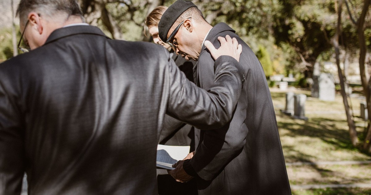 Men in formal attire comforting each other during an outdoor cemetery gathering.