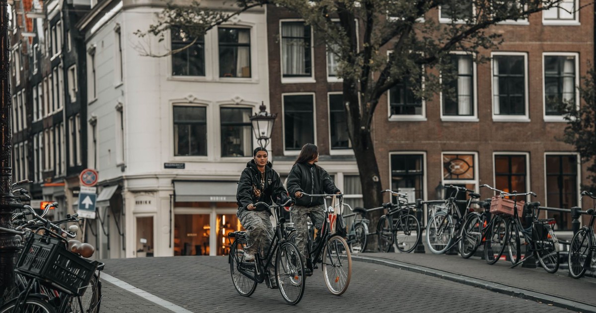 Two cyclists ride across a historic canal bridge in Amsterdam, surrounded by parked bicycles and traditional architecture.