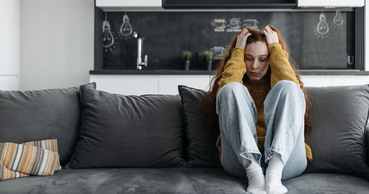 A woman in a yellow sweater sits on a couch, looking sad and pensive, indoors.