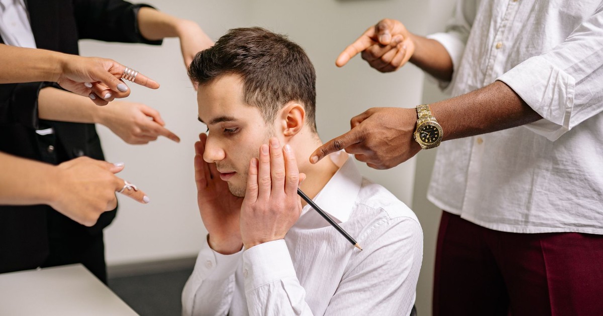 A stressed man at the office surrounded by pointing fingers, representing workplace bullying and stress.