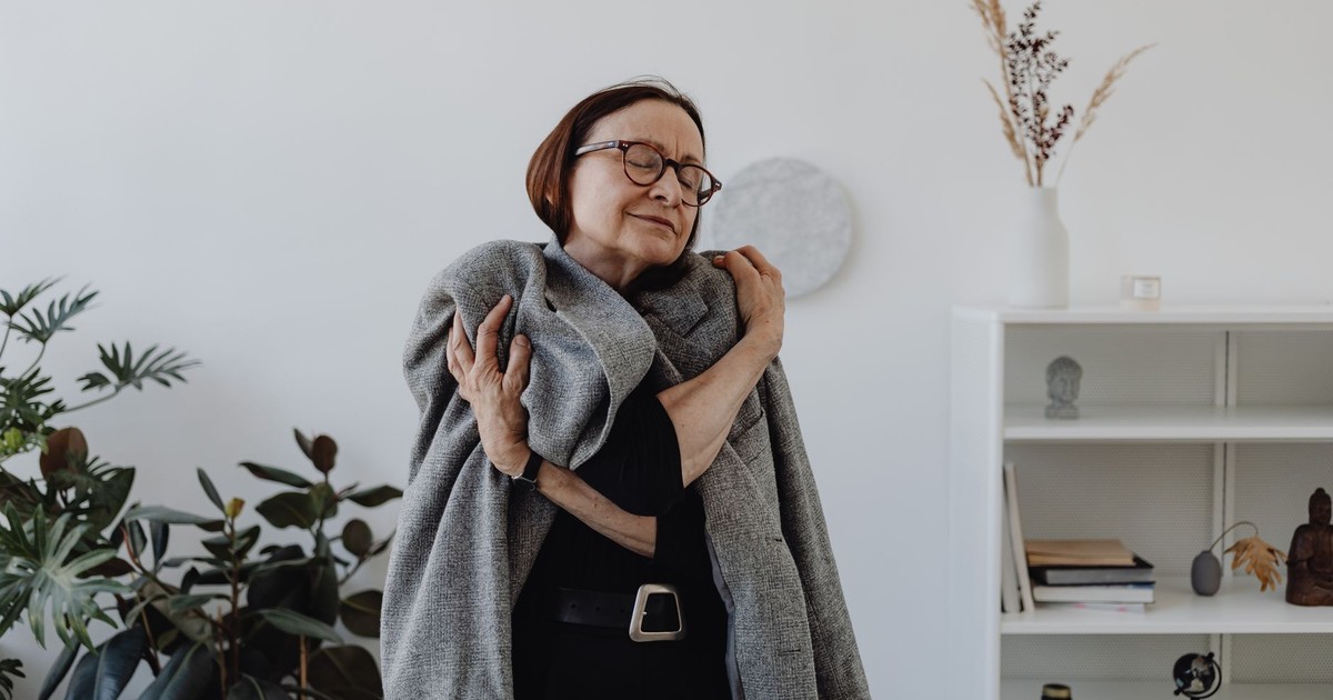 Smiling elderly woman in a black dress embracing a grey coat, conveying warmth.