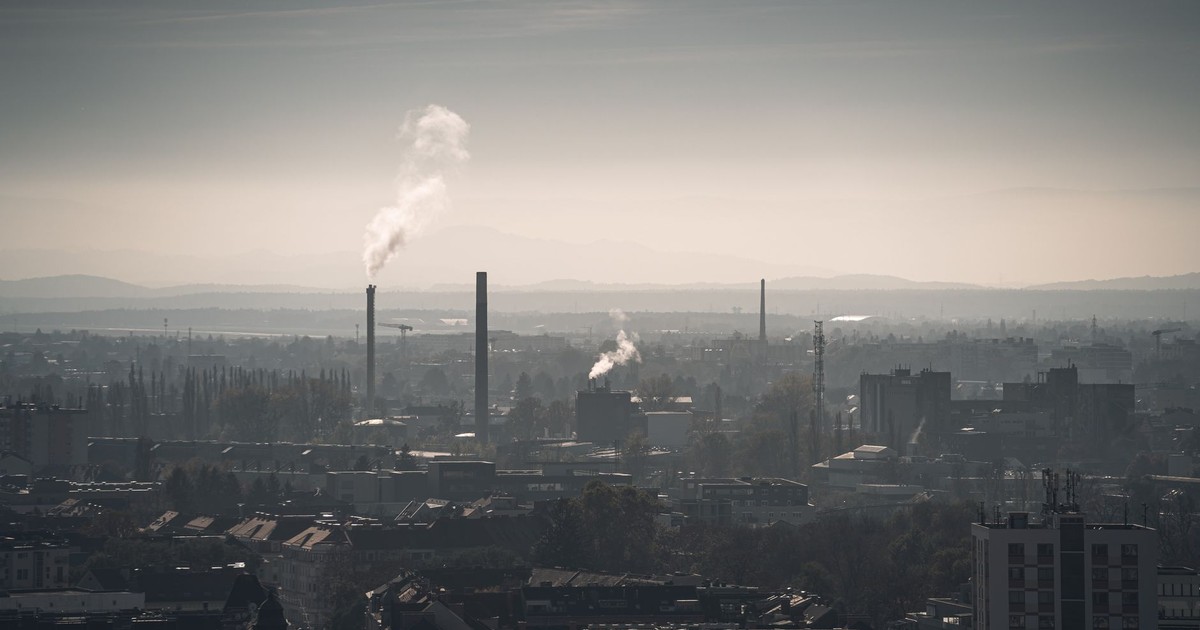 A cityscape with factory smokestacks emitting smoke, viewed at daybreak with a muted sky.