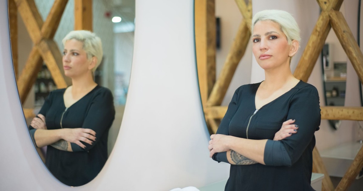 A professional hairdresser confidently stands in a modern salon with crossed arms, reflecting in a mirror.