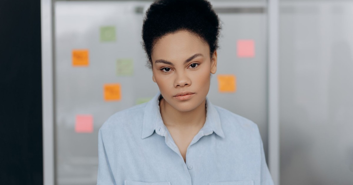 A woman wearing a blue shirt poses confidently in an office with sticky notes on the glass wall behind her.