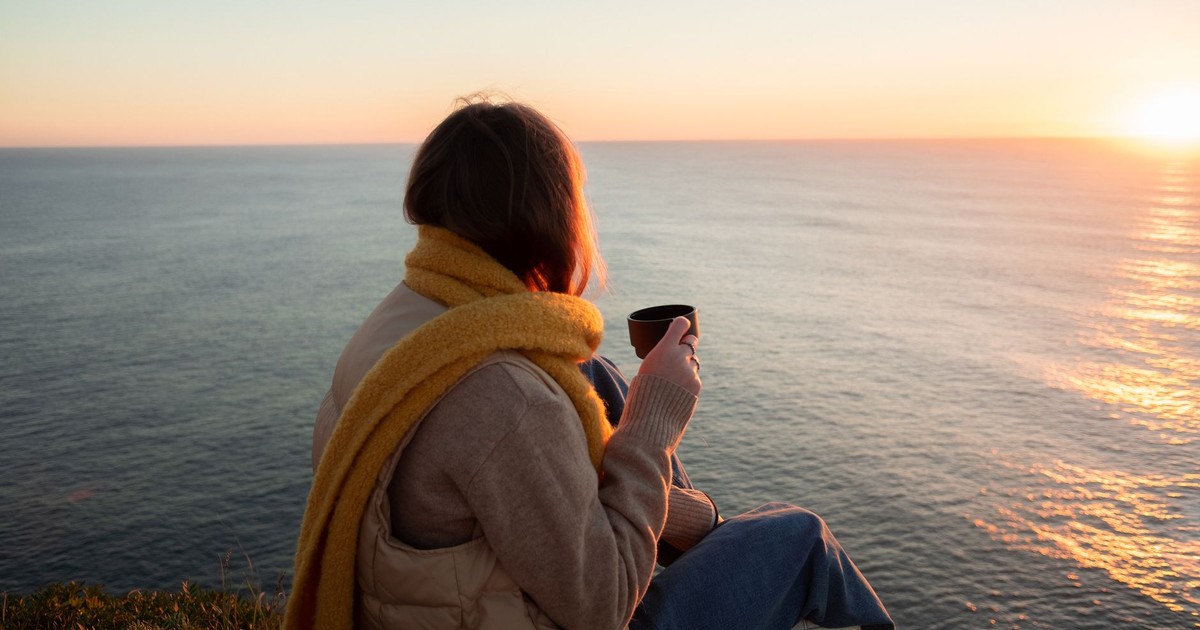 A woman in a yellow scarf enjoys a serene ocean view from a cliff at sunset, holding a mug.
