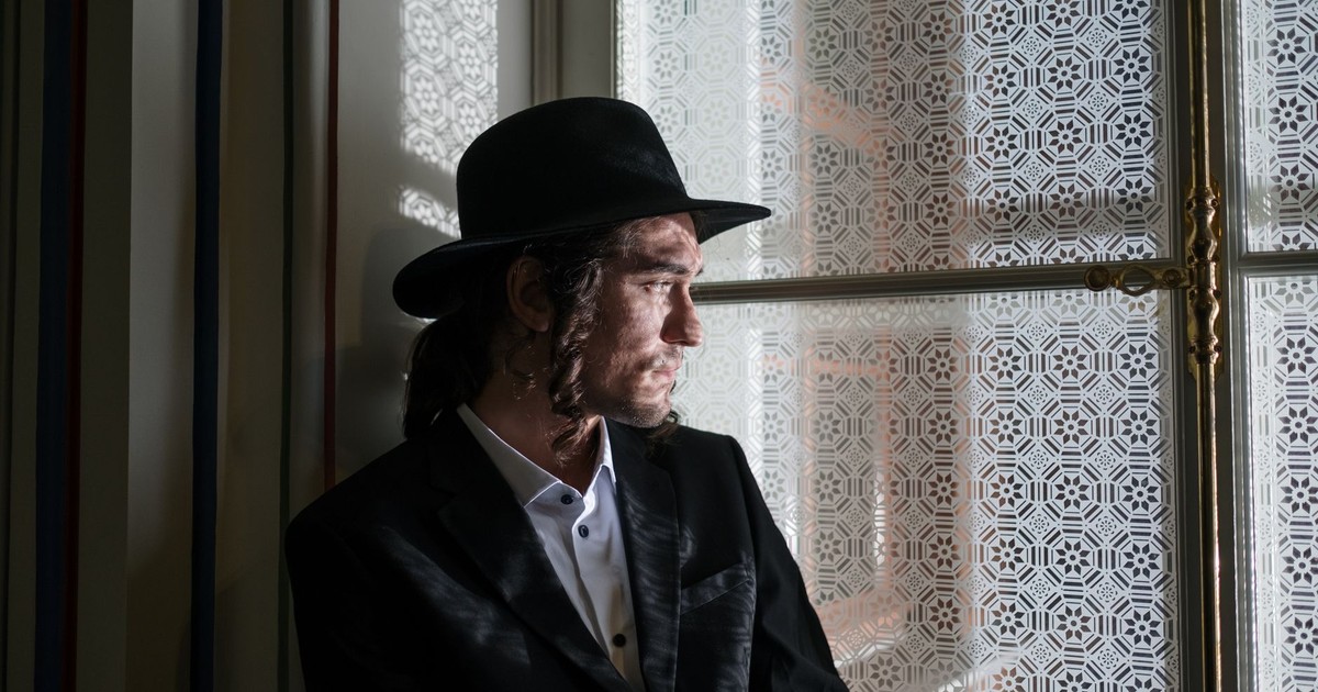 A thoughtful man in traditional Jewish clothing by a decorative window with sidelocks and a hat.