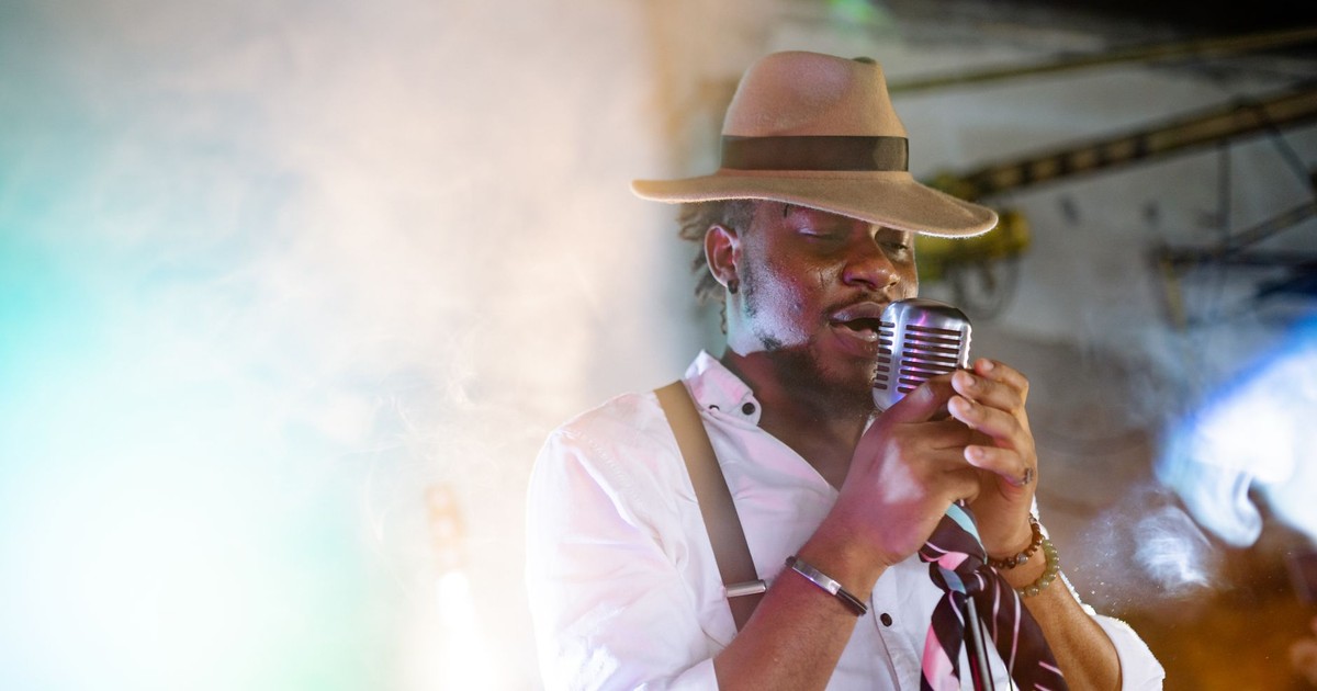 A stylish male singer performs passionately with a vintage microphone and hat on stage.
