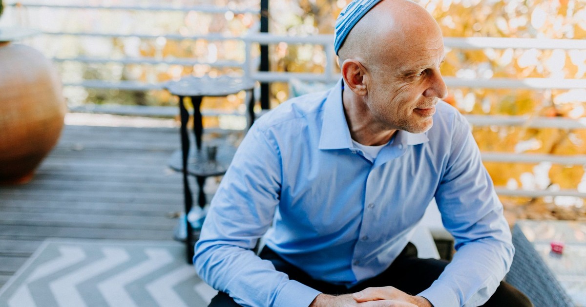 A cheerful man wearing a kippah sits outdoors in autumn, exuding warmth and joy.