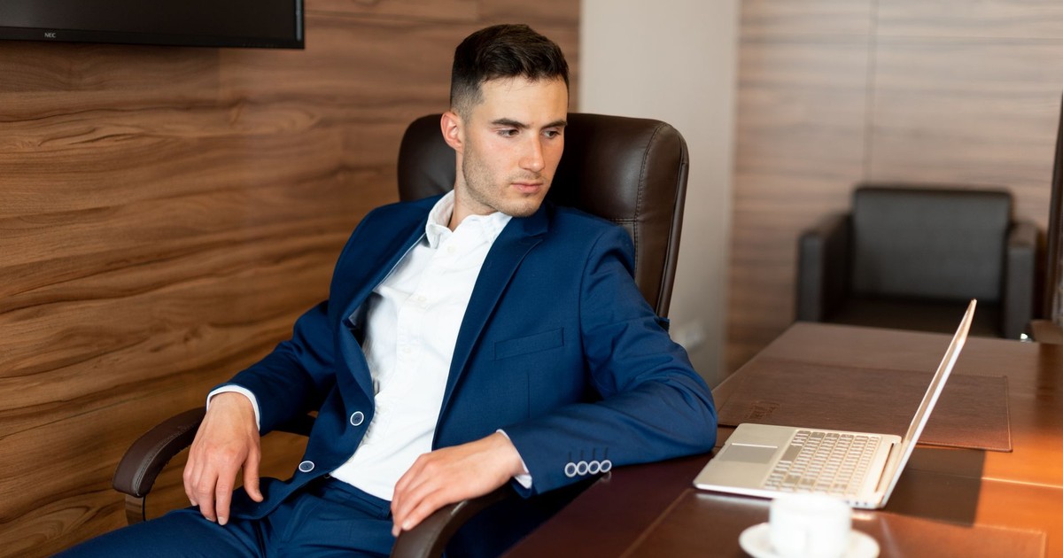 Confident businessman in blue suit seated at a desk with laptop, contemplating in office setting.